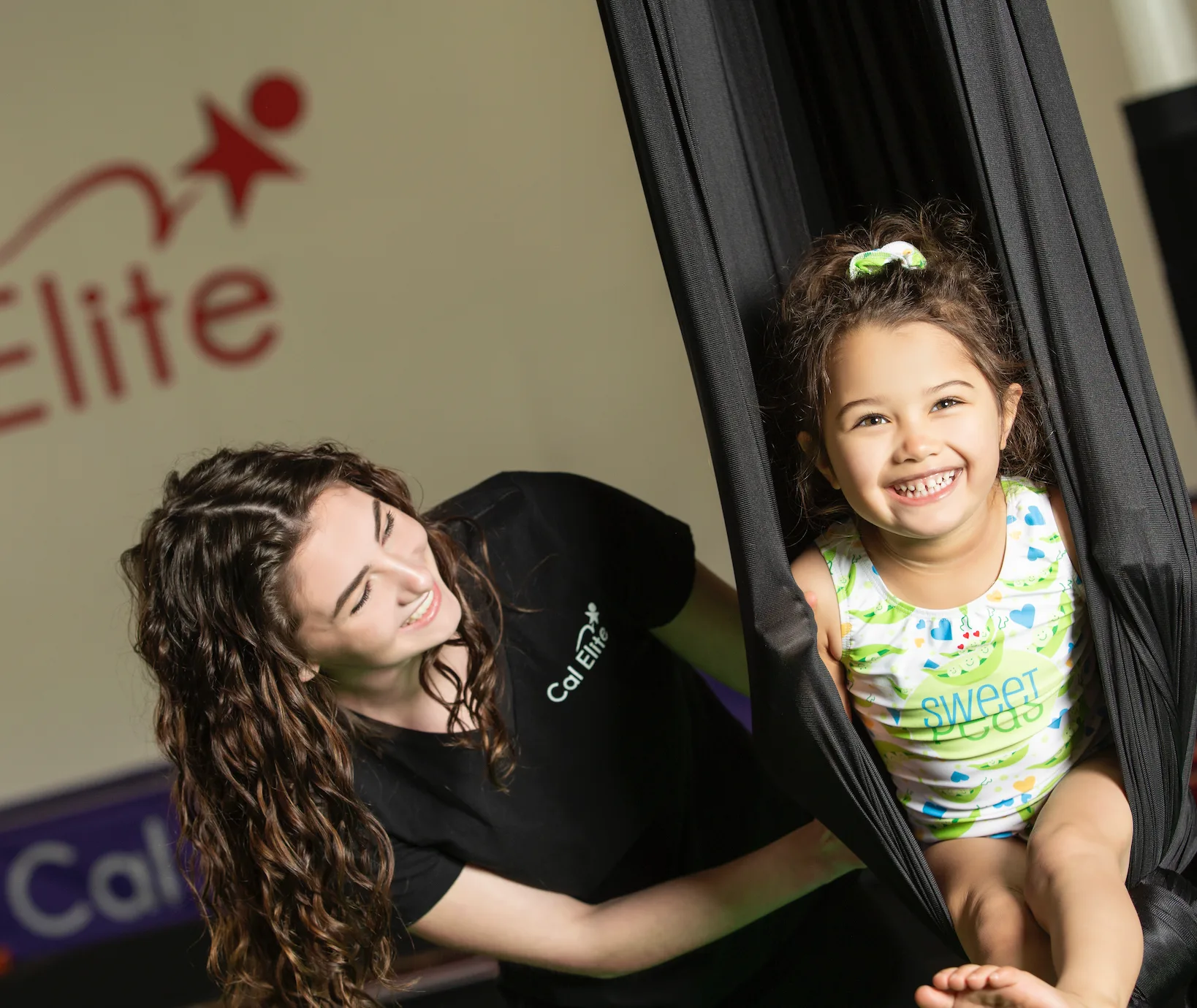 A Cal Elite coach helping a smiling young gymnast in an aerial hammock